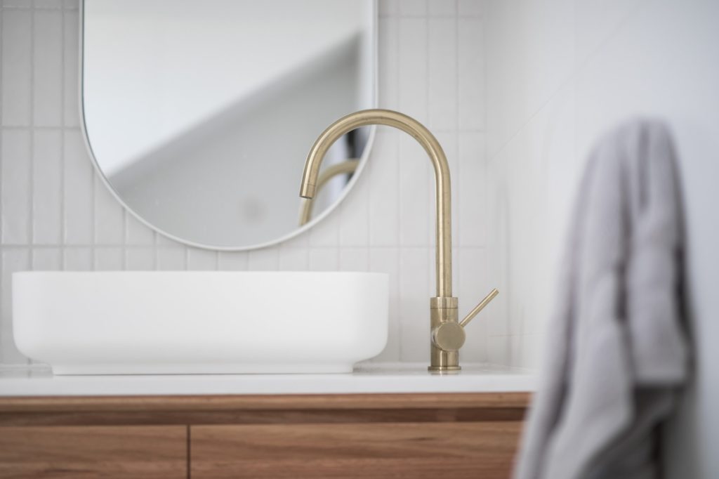 Brushed brass tap mixer on timber vanity with white basin bowl against white tiled wall in a new